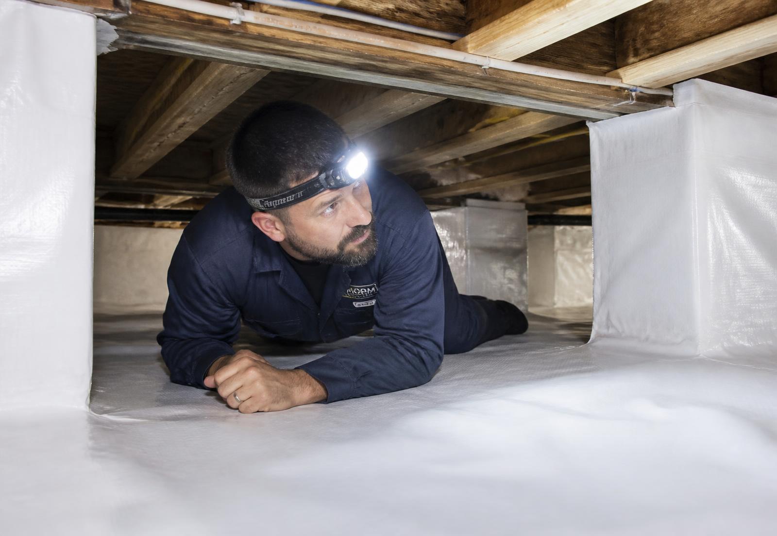Technician with a headlamp inspecting a sealed white crawl space beneath a home